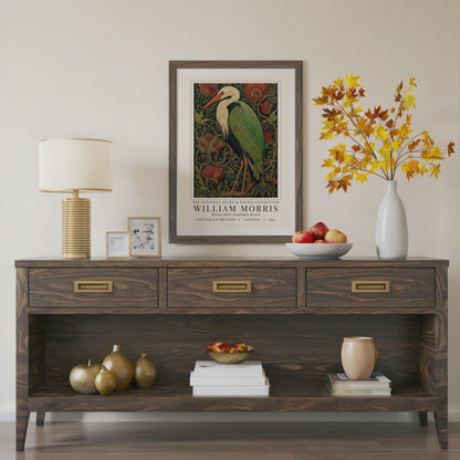 Wooden console table with decorative items including a lamp, framed artwork, and a vase with autumn leaves. William Morris inspired heron exhibition poster in a jewel-toned palette of forest green, antique crimson and old gold, featuring a grey heron amid ornate pimpernel blooms and curling vines — Victorian Arts and Crafts style, framed print.