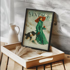 A framed vintage-style magazine cover print leaning on a wooden tray in a styled interior. The print shows a woman in a green coat and red hat walking a dog in the snow, with the word VINTAGE at the top.