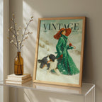 A framed vintage-style art print leaning on a glass console table in a styled interior. The print shows a woman in a green coat and red hat walking a black-and-white dog in the snow, with the word VINTAGE at the top.