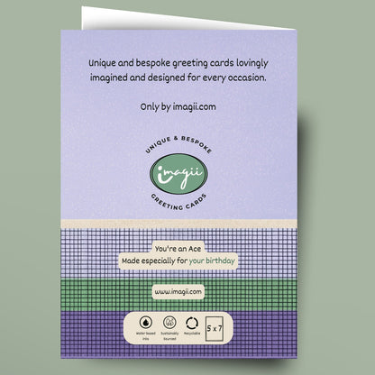 A greeting card shown against a muted sage green background. The card features a lavender top section with black text, a green circular logo reading 'Unique Bespoke Greeting Cards', and a patterned lower section with purple, green, and white plaid bands and a small white label area.