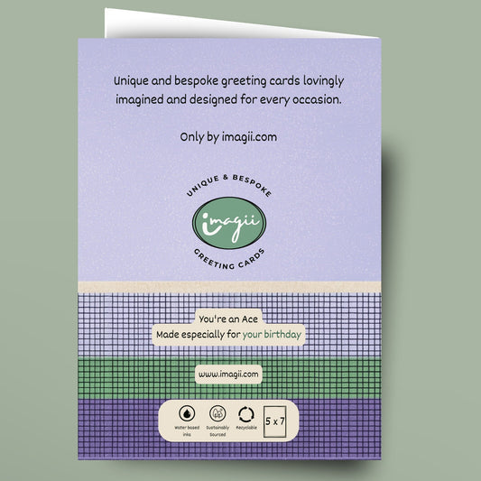 A greeting card shown against a muted sage green background. The card features a lavender top section with black text, a green circular logo reading 'Unique Bespoke Greeting Cards', and a patterned lower section with purple, green, and white plaid bands and a small white label area.