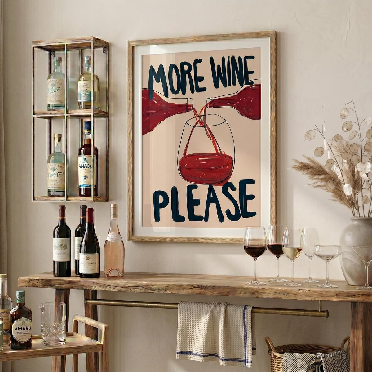 A framed wall art print with the text 'MORE WINE PLEASE' and an illustration of red wine pouring into a glass, displayed in a styled wine-themed room above a wooden console table with wine bottles, glasses, and dried flowers.