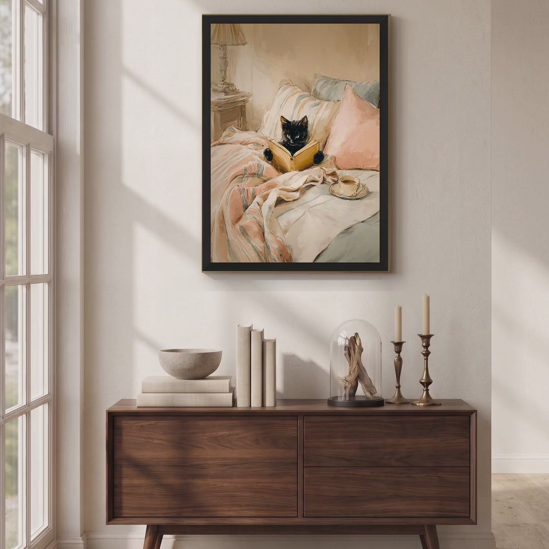 Video of a black-framed print of a black cat reading a book in bed with blush pink bedding, hung above a walnut sideboard in a sunlit room.