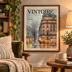 Wood-framed Vintoire Rainy Paris print hanging above a green velvet armchair and round side table in a cosy sitting area with trailing plants and a lamp.